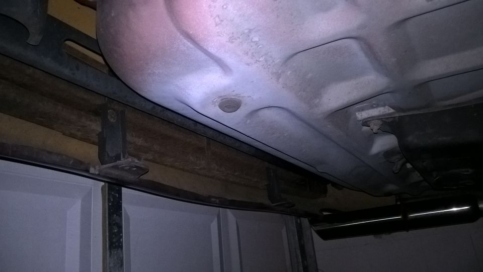 Underside view of a vehicle, showing the exhaust and part of the fuel tank. Garage door visible below