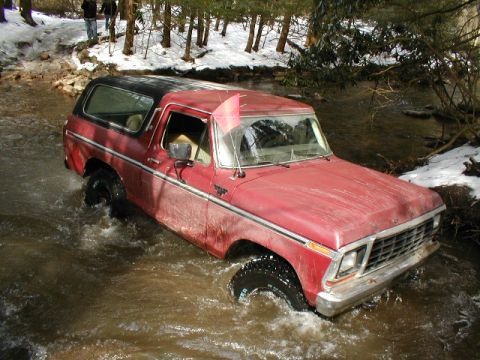 1979 Ford Bronco II - This red SUV is navigating a shallow stream in a wooded area with snow on the ground.