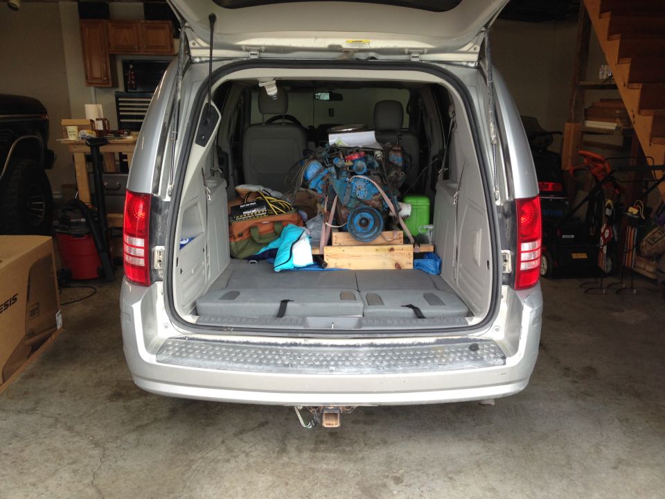 Silver minivan rear view with open trunk containing a blue engine on a wooden stand