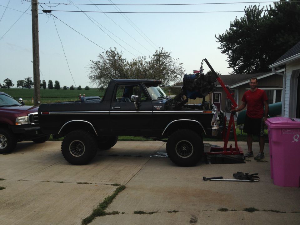 Black 1978 Ford Bronco with hood open, engine being lifted out by an engine hoist. Another vehicle is parked to the left
