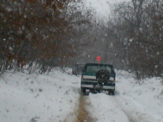 Two SUVs drive on a snow-covered road with trees on either side. One SUV has a spare tire mounted on the back and a red flag on top