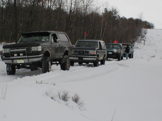 Lifted 1990s Ford Bronco leading a line of parked 4x4s on a snowy trail