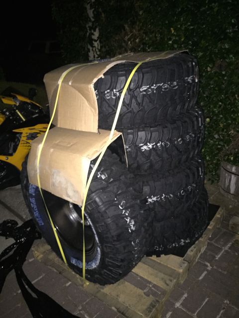1989 Ford Bronco - A stack of four off-road tires is packaged on a pallet, with a yellow motorcycle visible in the background.