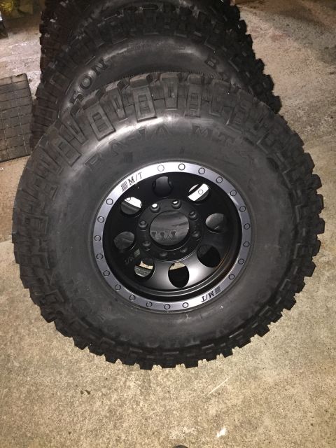 1989 Ford Bronco - Four off-road tires and black rims are stacked on a concrete floor.