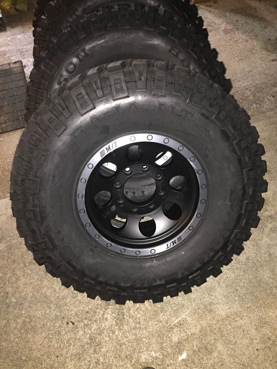 Stack of four off-road tires with black rims, some text visible on sidewalls