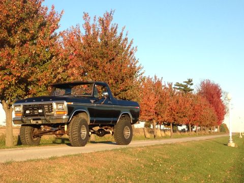 1979 Ford Bronco - A black 1979 Ford Bronco is parked on a dirt road lined with autumn trees, its lifted suspension and off-road tires.