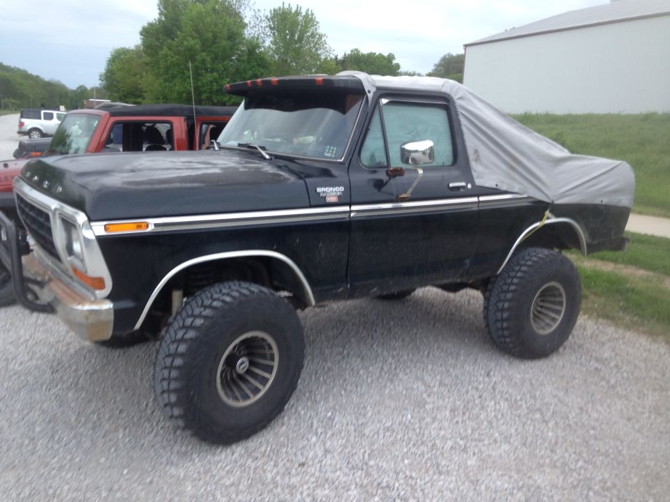 Black Ford Bronco with a grey tarp over the rear, lifted with large tires. Another red SUV and white truck are parked behind it