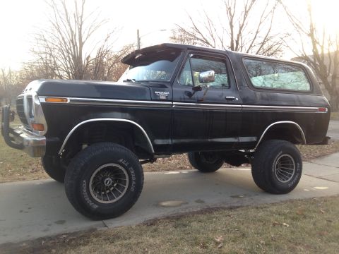 1979 Ford Bronco - This lifted black Ford Bronco is parked on a paved surface with trees in the background.
