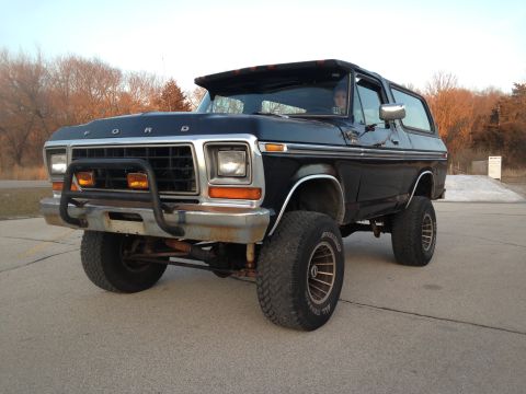1979 Ford Bronco - This lifted black Ford Bronco is parked on pavement with trees in the background.