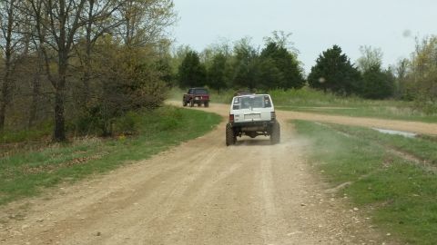 1979 Ford Bronco - Two vehicles drive down a dusty dirt road surrounded by trees and grass.