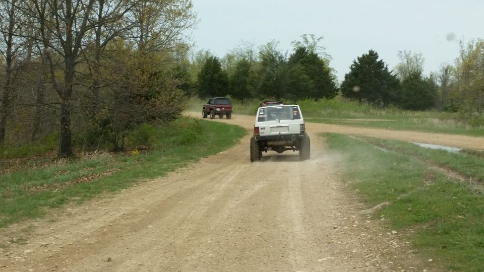 White SUV with large tires kicking up dust on a dirt road, followed by a red pickup truck
