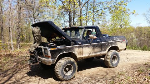 1979 Ford Bronco - Two people work on a dirty 1978 Ford Bronco in a dirt clearing surrounded by trees.