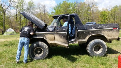1979 Ford Bronco - Two people attend to the engine of a muddy black Ford Bronco in an outdoor setting.