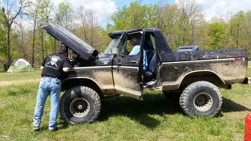 Black 1978 Ford Bronco with hood open, two people working on engine, parked in grassy field with tent in background