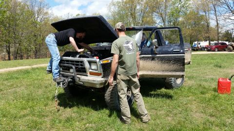 1979 Ford Bronco - Two men attend to the engine of a mud-covered Ford Bronco, with other vehicles visible in the distance.