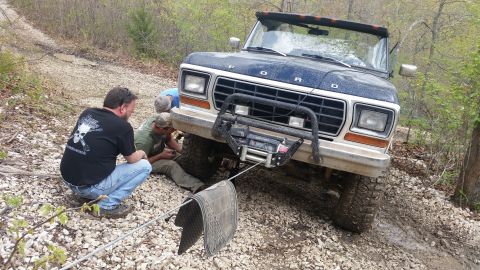 1979 Ford Bronco - Three men work on a dark blue 1979 Ford Bronco equipped with a front winch while it is stuck on a wooded, gravelly trail.