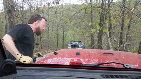 1979 Ford Bronco - Two off-road vehicles are on a dirt path in the woods, with a man observing the scene.