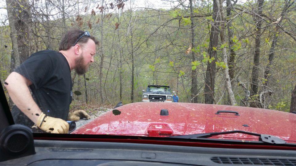 Man in black shirt and gloves looks at a dark-colored SUV ahead on a dirt road in a wooded area. Red vehicle hood in foreground