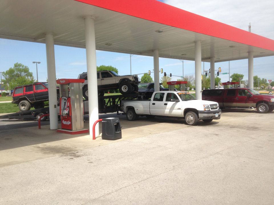 White dually truck hauling a lifted Jeep on a trailer, with another Jeep on a trailer and a red pickup truck at a gas station