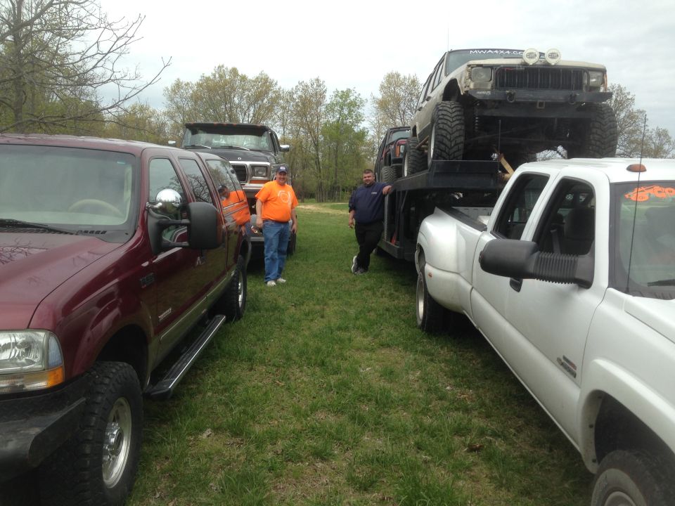 Several trucks are parked on grass, one is a flatbed hauling a Jeep. Two men are standing around