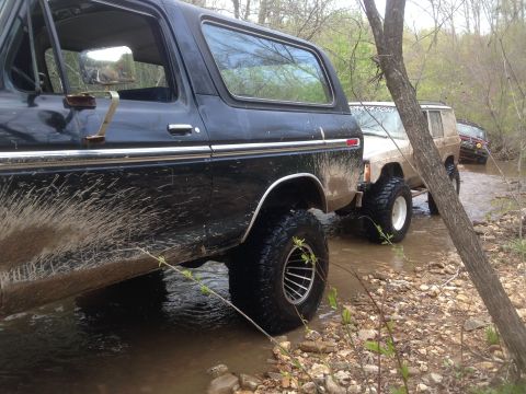 1979 Ford Bronco - Several off-road vehicles navigate a shallow stream surrounded by trees and muddy banks.