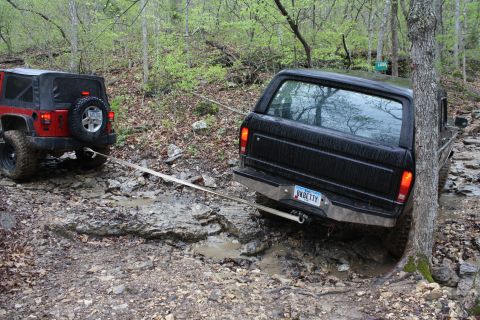 1979 Ford Bronco - Two off-road vehicles, a black Ford Bronco and a red Jeep, are stuck in the mud on a wooded trail.