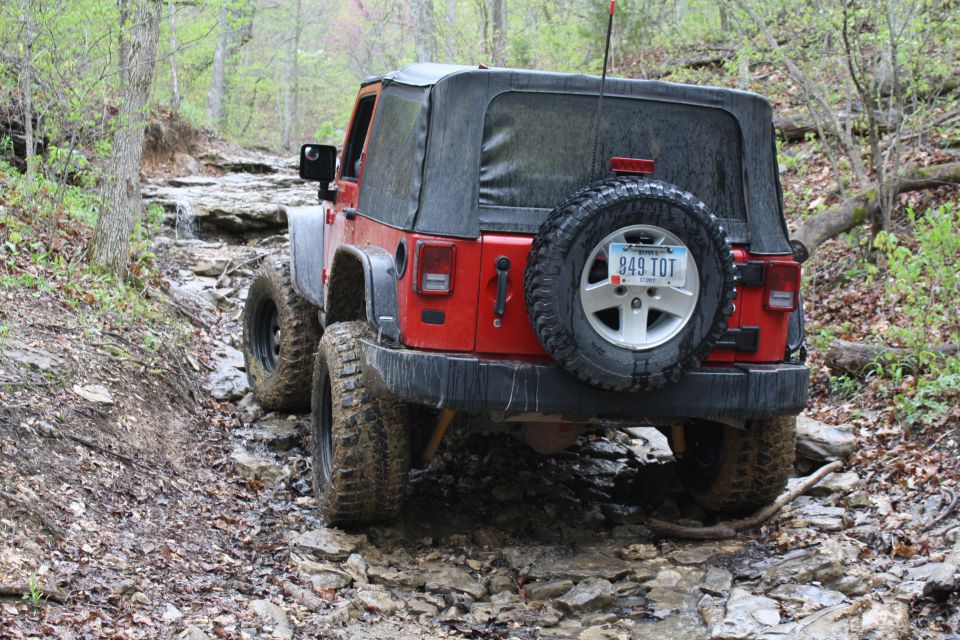 Red Jeep Wrangler with a soft top navigates a muddy, rocky trail from the rear