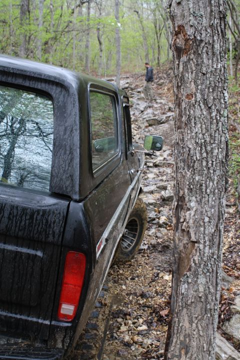 1979 Ford Bronco - A dark-colored SUV navigates a challenging, wet, and rocky path through a wooded area.