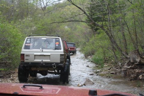 1979 Ford Bronco - A convoy of off-road vehicles navigates a water-filled path through a wooded area.
