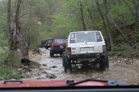 1979 Ford Bronco - Several Jeeps navigate a muddy, wooded path, kicking up water as they go.
