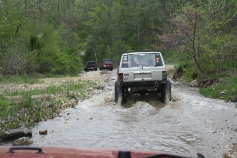 1979 Ford Bronco - Off-road vehicles navigate a water crossing on a dirt path surrounded by trees.