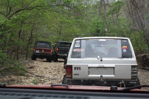 1979 Ford Bronco - A convoy of off-road vehicles navigates a wooded trail, kicking up dust on the rocky path.