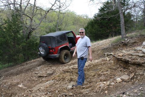 1979 Ford Bronco - A man poses with his red Jeep Wrangler on a muddy, off-road trail surrounded by trees.