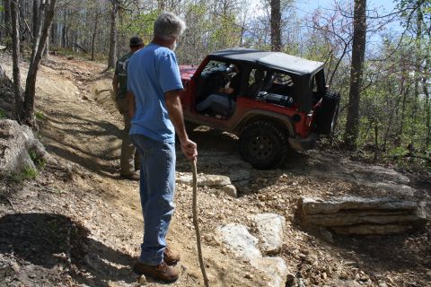 1979 Ford Bronco - A red Jeep Wrangler navigates a challenging rocky trail in a wooded area, with two onlookers observing the off-road adventure.