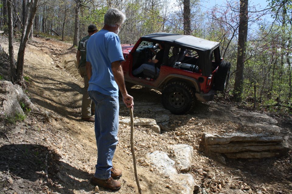 Red Jeep Wrangler with a soft top on a rocky, wooded trail, with two men standing nearby