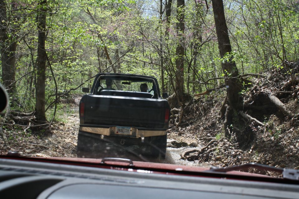 Black pickup truck driving on a muddy trail through trees. Another vehicle is visible in the foreground