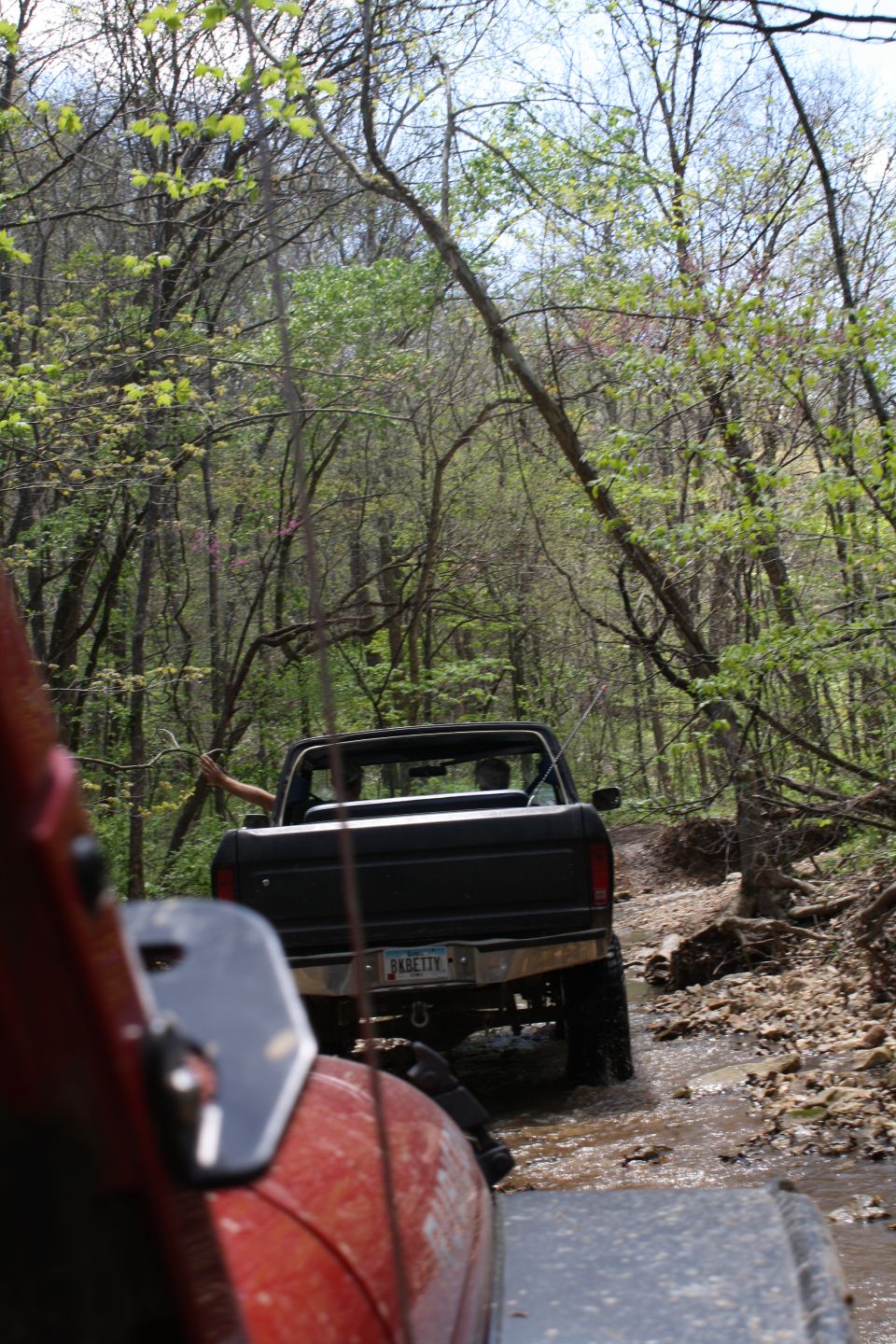 Black 1979 Ford Bronco driving on a muddy trail, seen from behind another vehicle