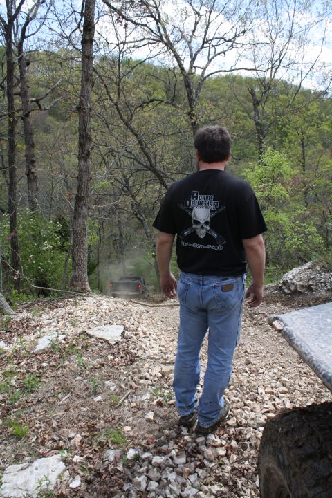 1979 Ford Bronco - A man in a black t-shirt and jeans watches an off-road vehicle navigate a dusty, wooded trail.
