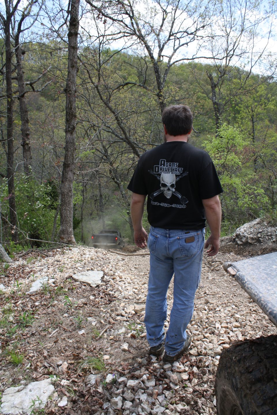 Dark off-road vehicle driving on a rocky, wooded trail, kicking up dust