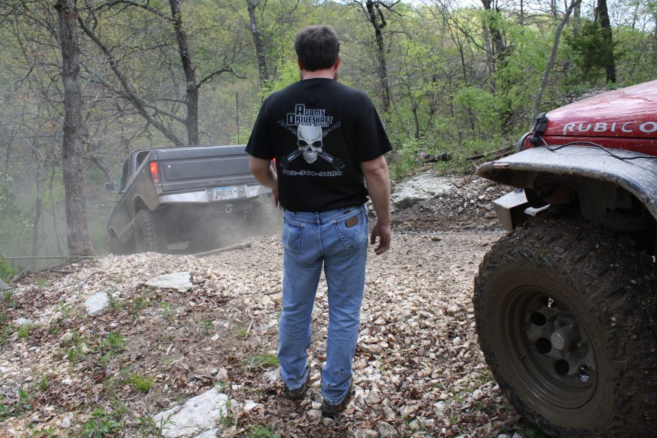 Man in jeans and t-shirt watches a pickup truck navigate a dusty, rocky off-road trail; a red Jeep is parked nearby