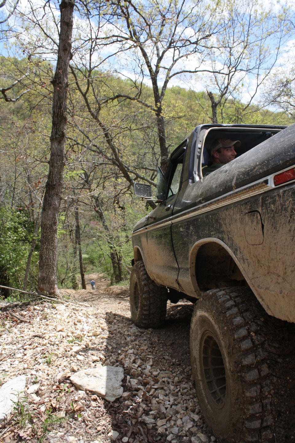 Mud-splattered dark green 1978 Ford Bronco, 3/4 front view, driving on a rocky, wooded trail
