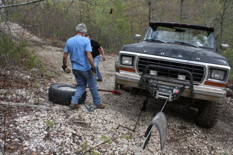 1979 Ford Bronco - A black 1979 Ford Bronco is being winched on a gravel path, with two men attending to a spare tire.