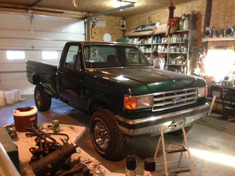 1989 Ford F-250 - A green Ford pickup truck is parked inside a cluttered garage with shelves of supplies in the background.