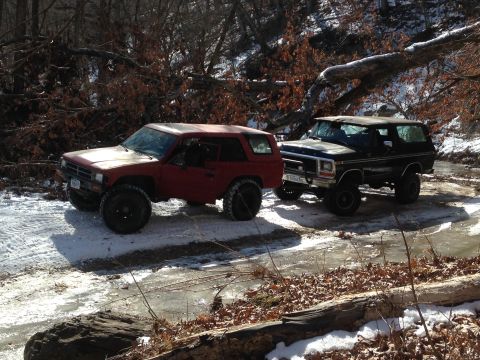 1979 Ford Bronco - Two SUVs are parked on a slippery, snow-covered road surrounded by bare trees and fallen logs.