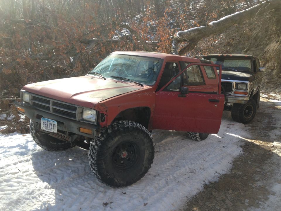 Red Toyota pickup truck with open driver's door, large tires, and a black Ford Bronco behind it in a snowy, wooded area