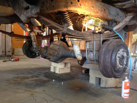 1979 Ford Bronco - Underside of a truck on cinder-block jack stands showing rear leaf springs, axle, drum brake and a work light.