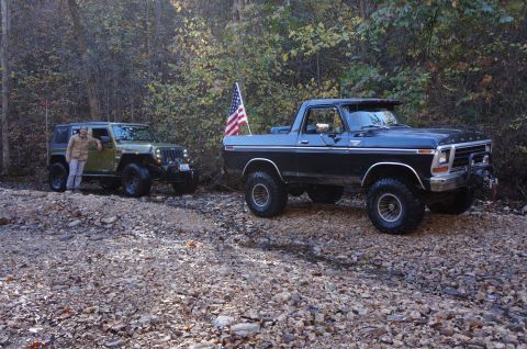1979 Ford Bronco - Two off-road vehicles, a 1979 Ford Bronco and a Jeep Wrangler, are parked on a rocky path in a wooded area.