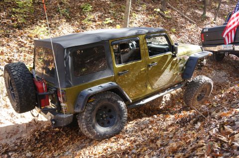1979 Ford Bronco - A Jeep Wrangler navigates a wooded trail with fallen leaves, while another vehicle is visible behind it.