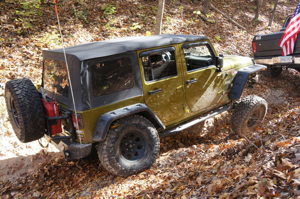 Olive green Jeep Wrangler with a soft top, spare tire on the rear, and off-road tires on a dirt trail