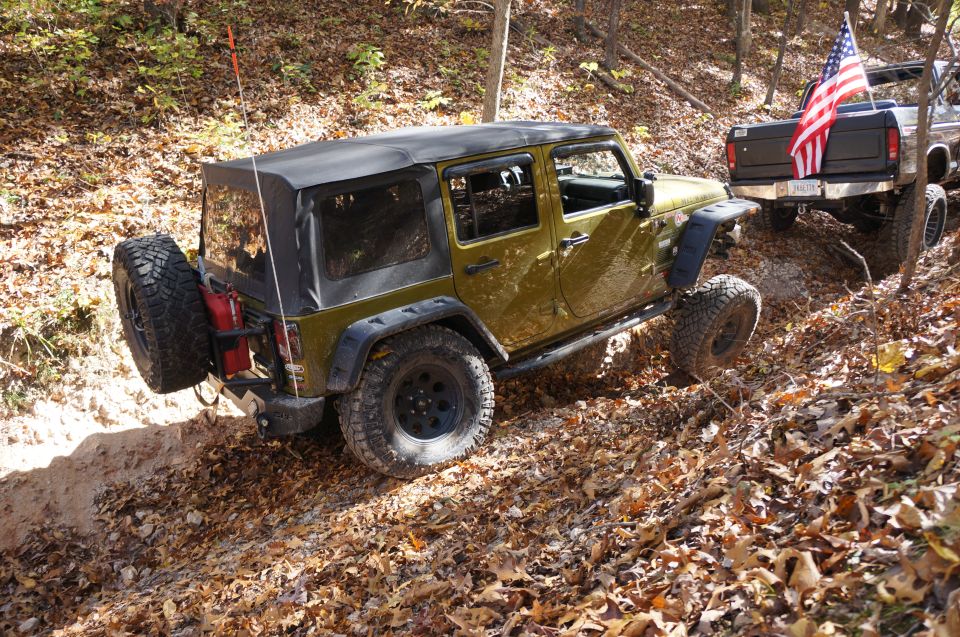 Olive green Jeep Wrangler and a pickup truck with an American flag drive on a leaf-covered dirt trail